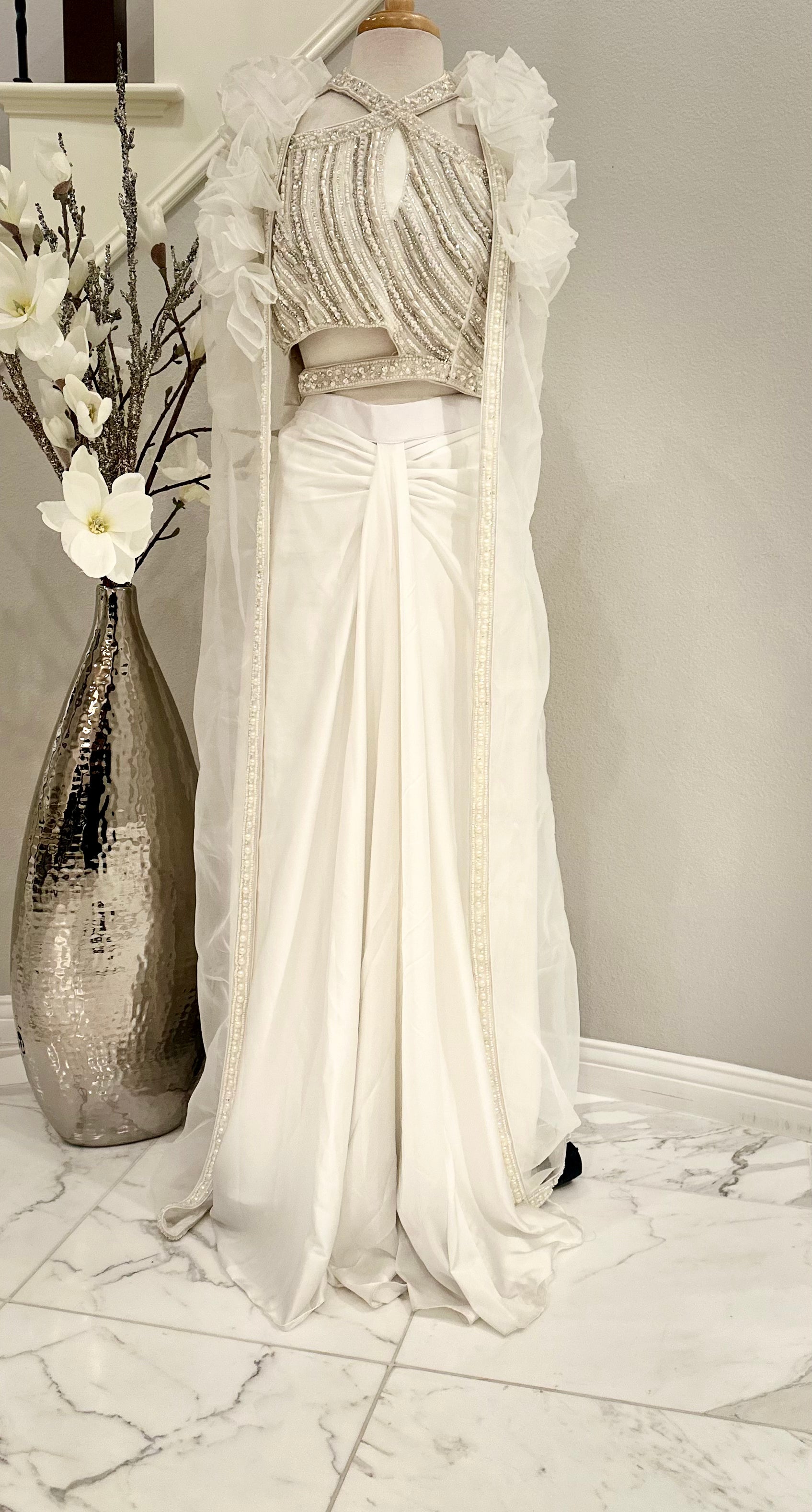 Beautifully crafted 3 piece shrug set in pure silk with pearl embellishment on the asymmetrical crop-top. Paired with long open shrug  in soft organza with pearl lace on the open edges. Paired with a plain white pleated front skirt

Size available 38 with margin.